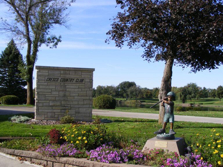 Cresco Country Club stone sign with a bronze status of a child golfing and flowers in front of it.