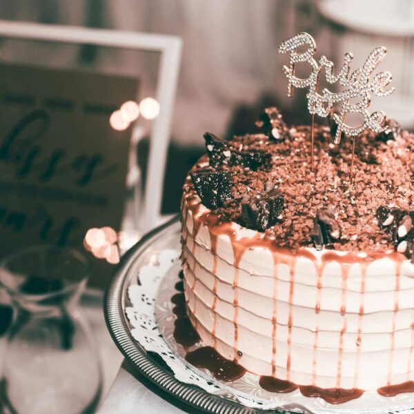 Layered cake with caramel drizzle and chocolate decorations on a white lace doily, topped with a "Bride" sign. Nearby, a framed sign is slightly blurred in the background.