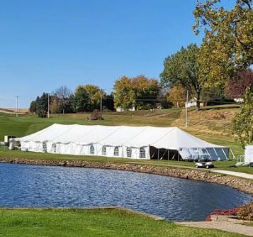 A large white tent is set up on a grassy area beside a pond, with trees and a clear blue sky in the background.