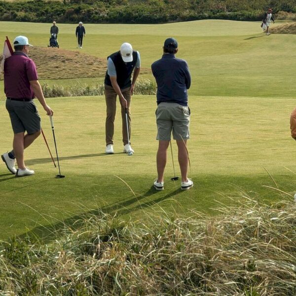 Four golfers on a green, with one putting while the others watch. Golf clubs are on the ground, and other people are in the background on the course.