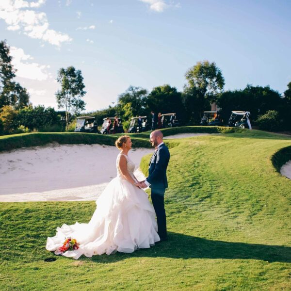 A bride and groom stand holding hands on a grass hill with sand bunkers, surrounded by trees and blue sky.